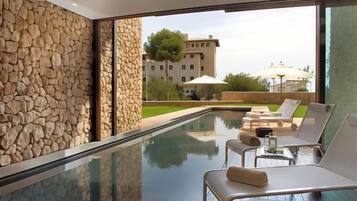 Un área de piscina interior moderna con paredes de piedra, sillas de descanso y vista a un edificio al fondo.