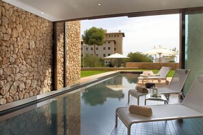 A modern indoor pool area with stone walls, lounge chairs, and a view of a building in the background. - Hotel Hospes Maricel & Spa (Ca's Catala)