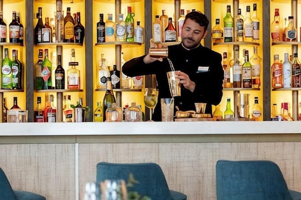 A bar area with shelves of various liquor bottles, a bartender preparing a drink, and seating in the foreground.