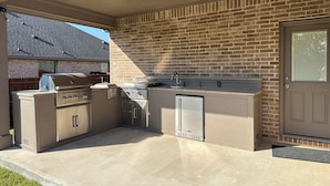 An outdoor kitchen with a grill, sink, and storage cabinets, adjacent to a brick wall and a door.