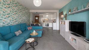 A modern living room with a sofa, a small table, and a TV. There is a kitchen in the background.