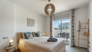 A bedroom with a bed, bedside lamp, decorative wall piece, and a view of the outside.