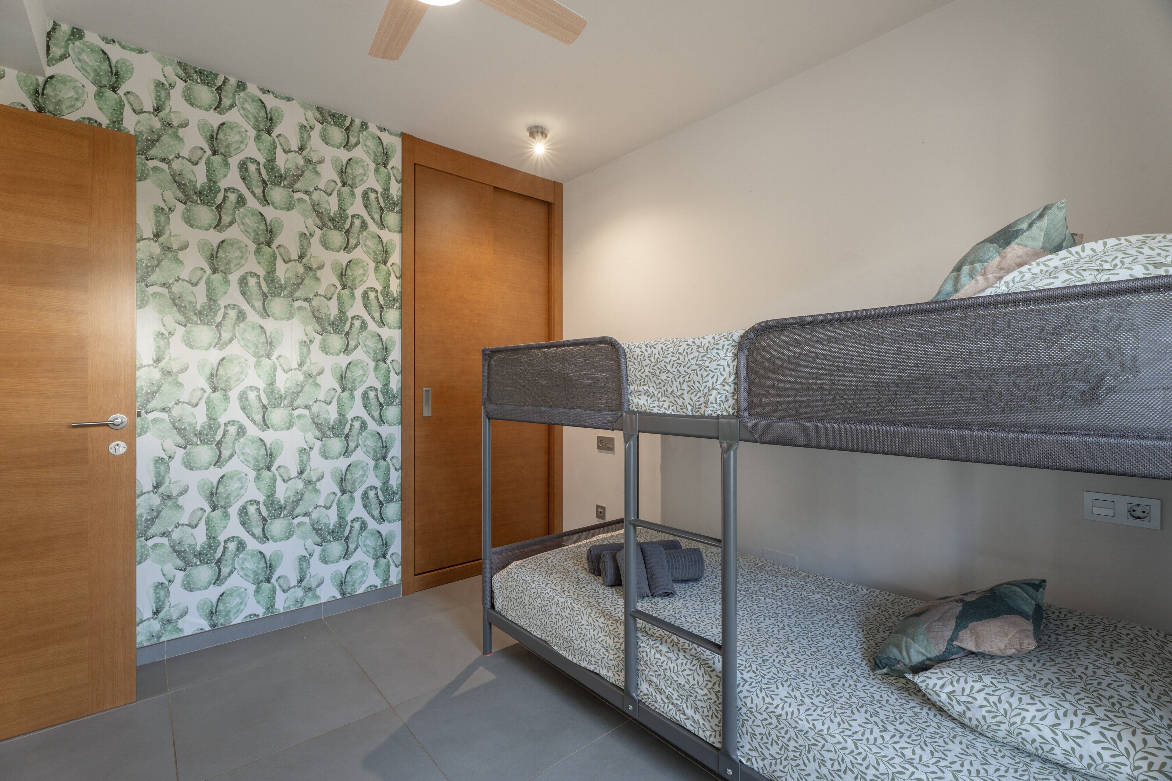 Uma cama beliche com colchão estampado, uma porta de madeira e papel de parede com tema de cactos.