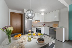Une cuisine moderne avec des meubles blancs, une table à manger agrémentée de fruits et de boissons, et des luminaires suspendus.