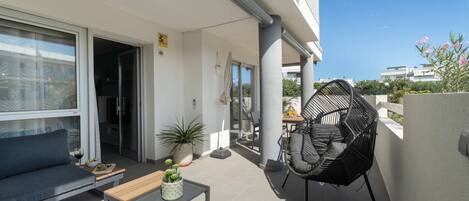A balcony with a sofa, a table with a plant, a chair, and a view of the surrounding buildings.