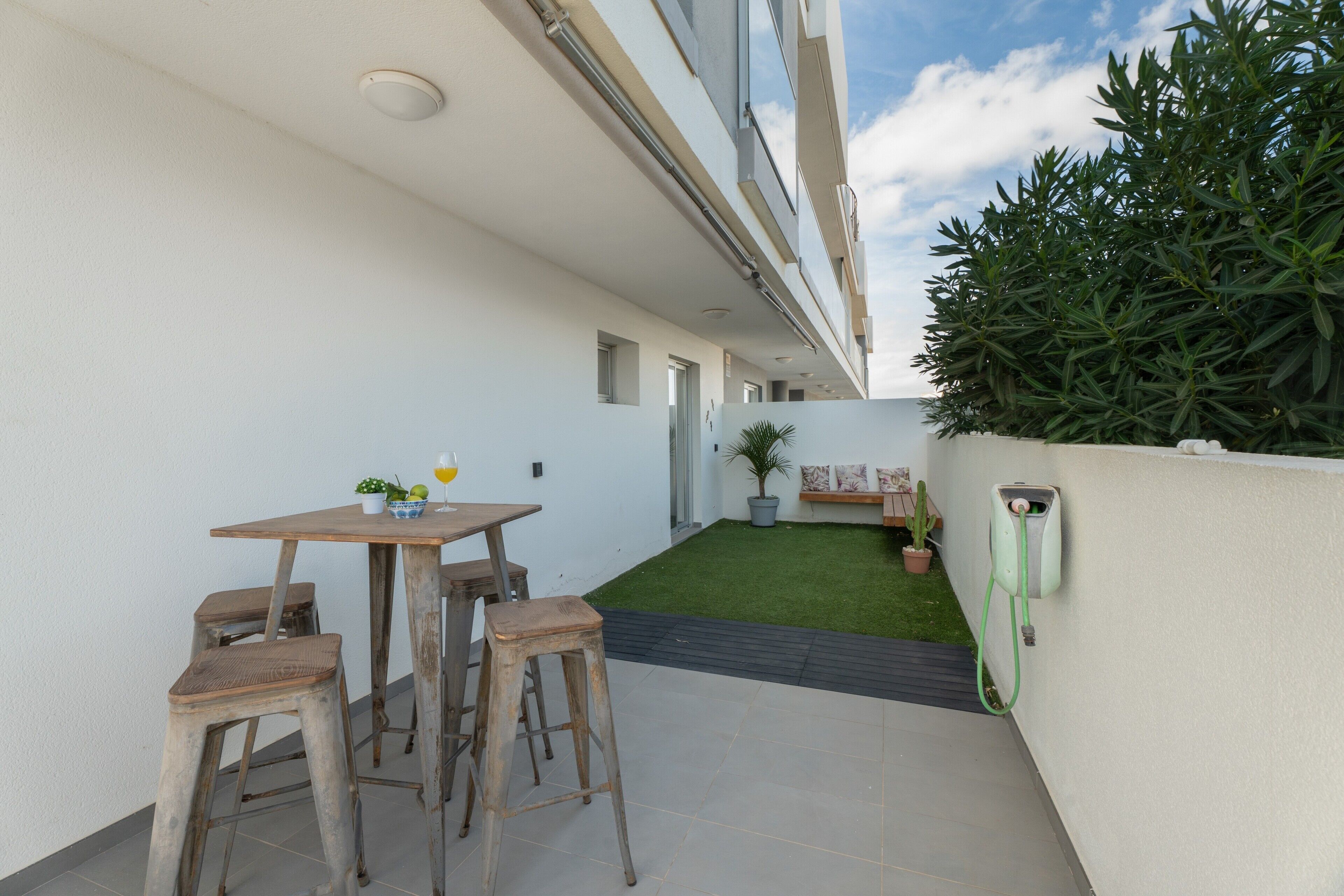 A patio with a table and stools, a small garden area with a potted plant, and a green wall-mounted device.