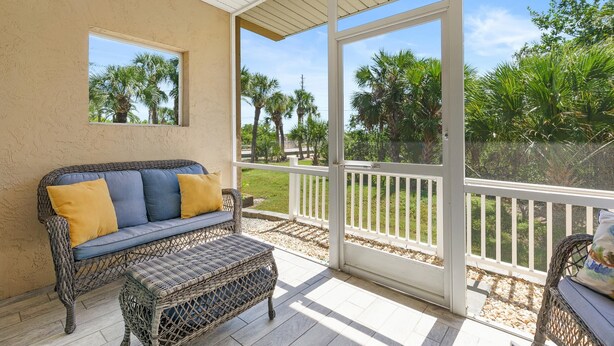A covered patio with a wicker sofa and ottoman, a view of palm trees, and a white railing.