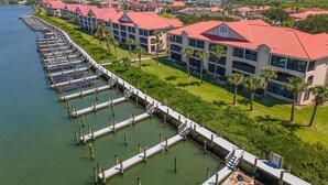 A marina with docked boats, a row of buildings with red roofs, and a grassy area with palm trees.