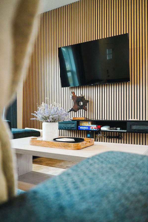 A modern living room with a flat-screen TV mounted on a wooden wall, a coffee table with a decorative vase, and a bookshelf with books and a small speaker.