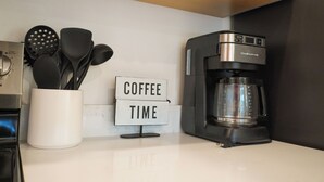 Un comptoir de cuisine avec une machine à café, une pancarte indiquant « PAUSE CAFÉ », et un récipient contenant des ustensiles de cuisine.