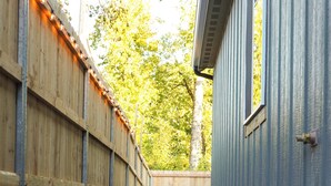 Un chemin mène à un bâtiment qui est bordé d’une clôture en bois d’un côté et d’un revêtement métallique de l’autre.