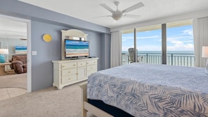 Een hotelkamer met een groot bed, een plafondventilator, een televisie op een dressoir en een balkon met uitzicht op de oceaan.