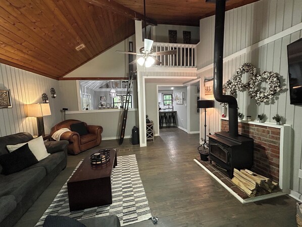 A spacious living room with a high ceiling, a fireplace, a staircase leading to an upper level, and a view into the kitchen.