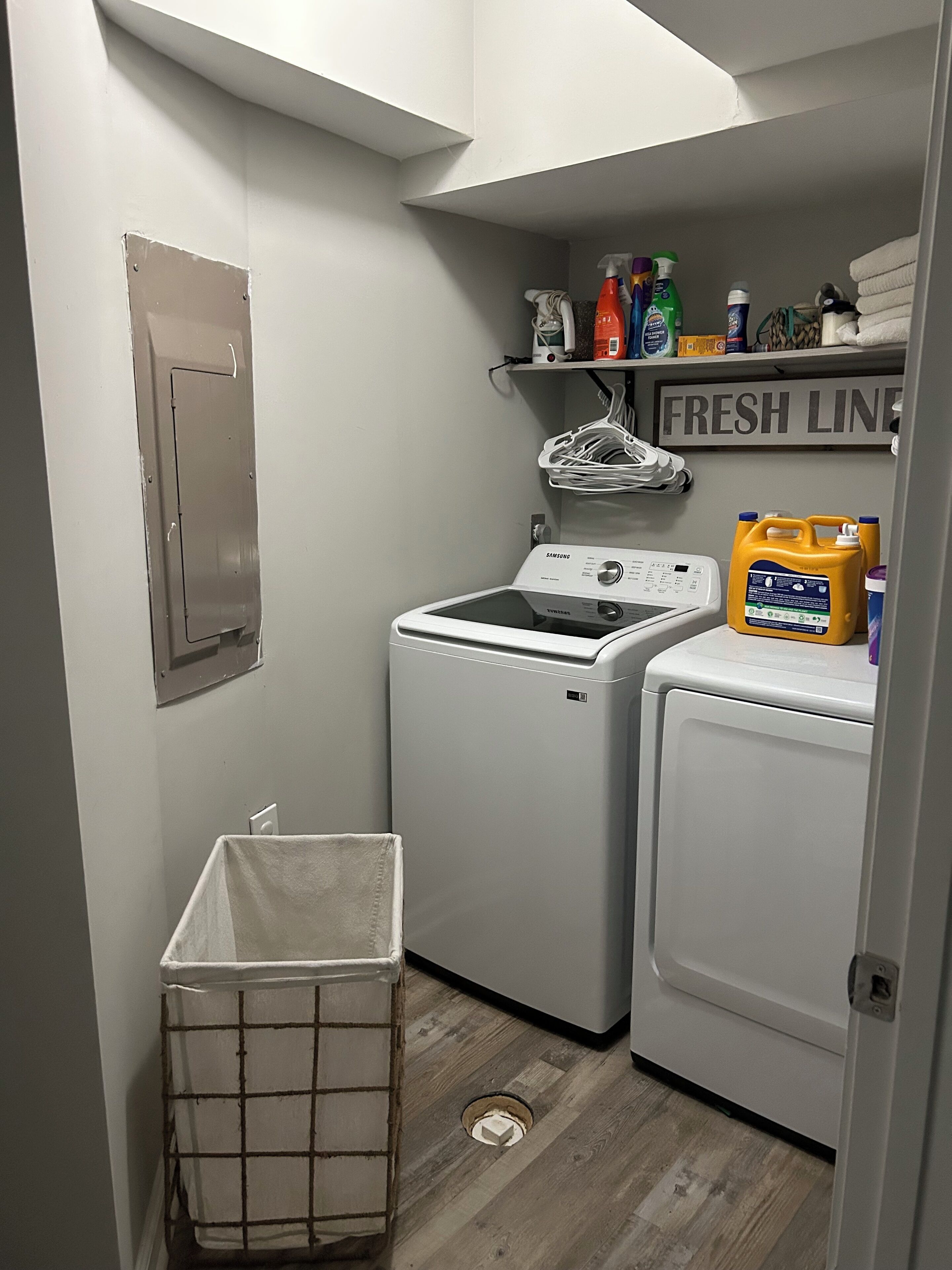 A compact laundry room with two washing machines, a drying machine, a shelf with cleaning supplies, and a metal laundry basket.