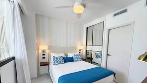 Une chambre à coucher comprenant un lit, des tables de chevet, un miroir, un ventilateur de plafond et une porte.