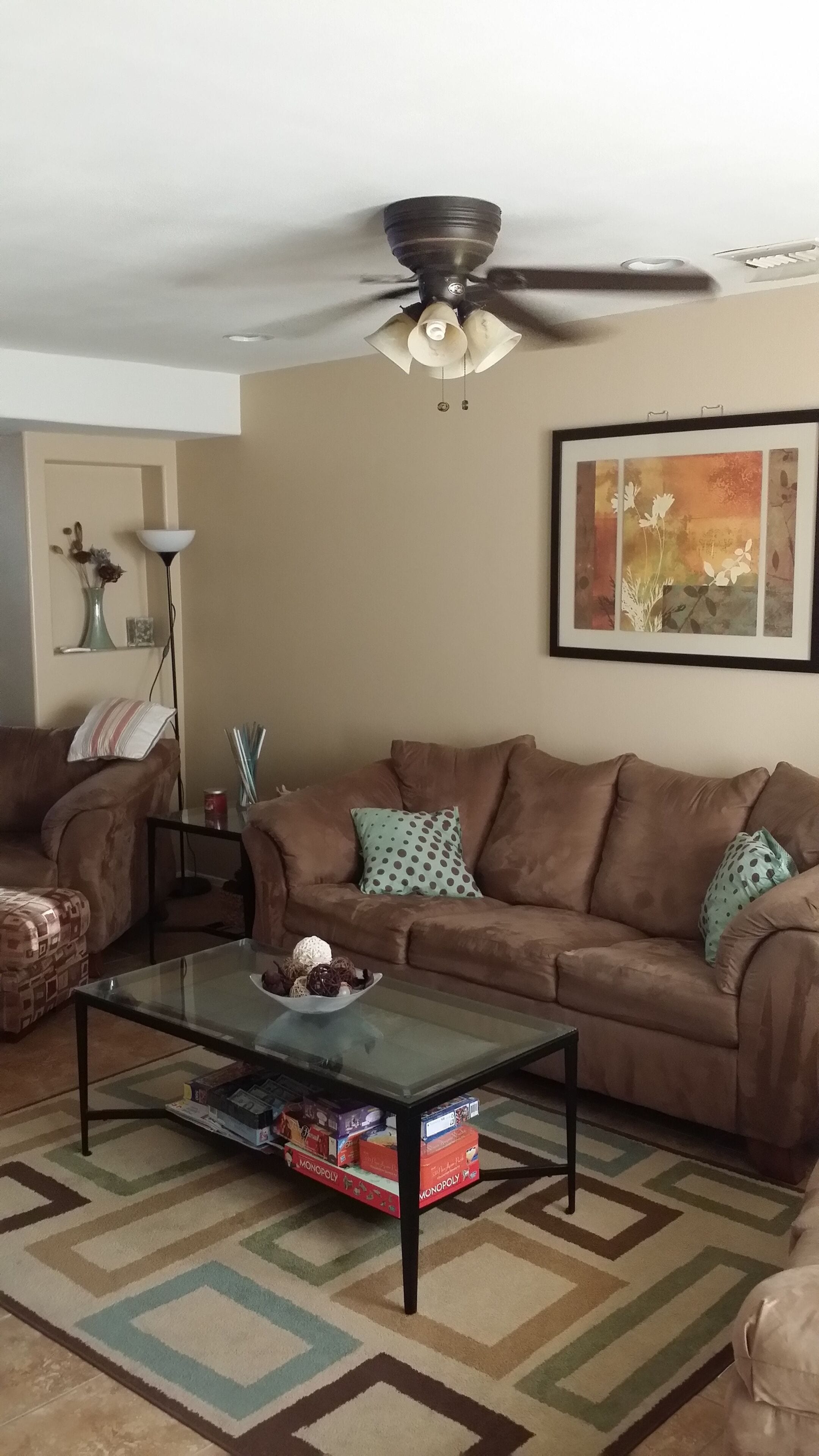 A living room with a brown sofa, a glass coffee table, a ceiling fan, a framed artwork, and a patterned rug.