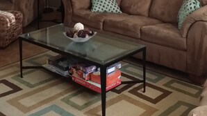 Ein Wohnzimmer mit braunem Sofa, einem Glastisch, einem Deckenventilator, einem gerahmten Kunstwerk und einem gemusterten Teppich.