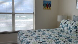 Une chambre avec un grand lit, une fenêtre avec des stores, un tableau représentant des planches de surf et une lampe de chevet.