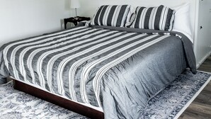 A neatly made bed with a striped bedspread and pillows, a bedside table with a lamp, and a patterned rug on a wooden floor. - White House on Market (Hermann)