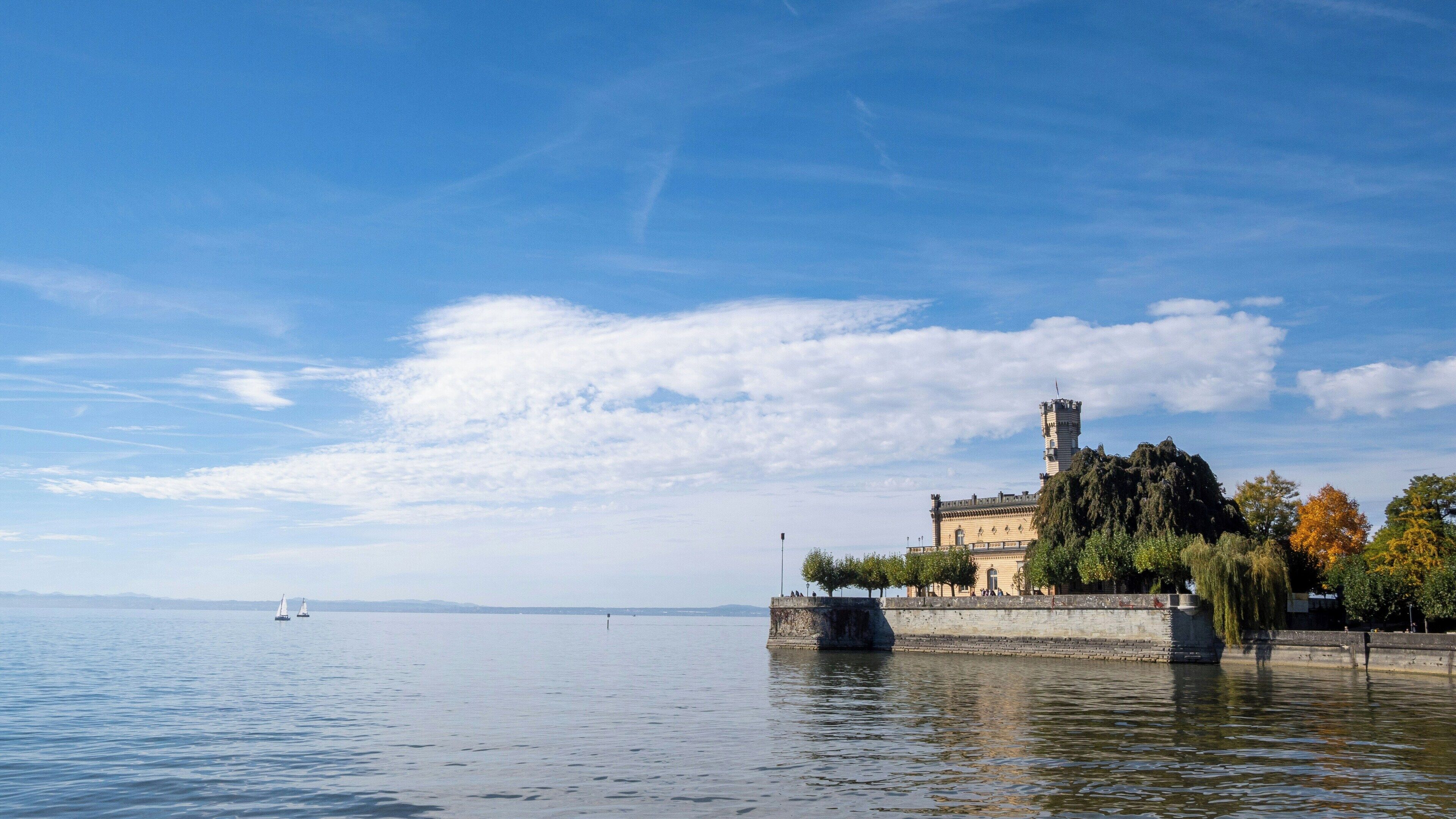 A historic building with a tower, situated by a calm body of water, with a clear sky above.