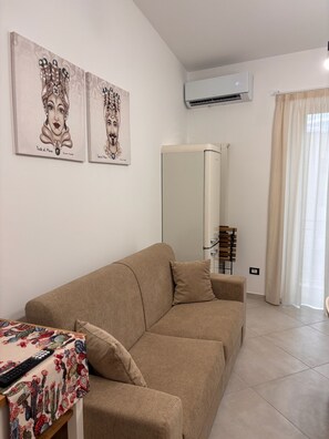 Een woonkamer met een beige bank, een bijzettafeltje met een kleurrijke tafelkleed en twee kunstwerken aan de muur.