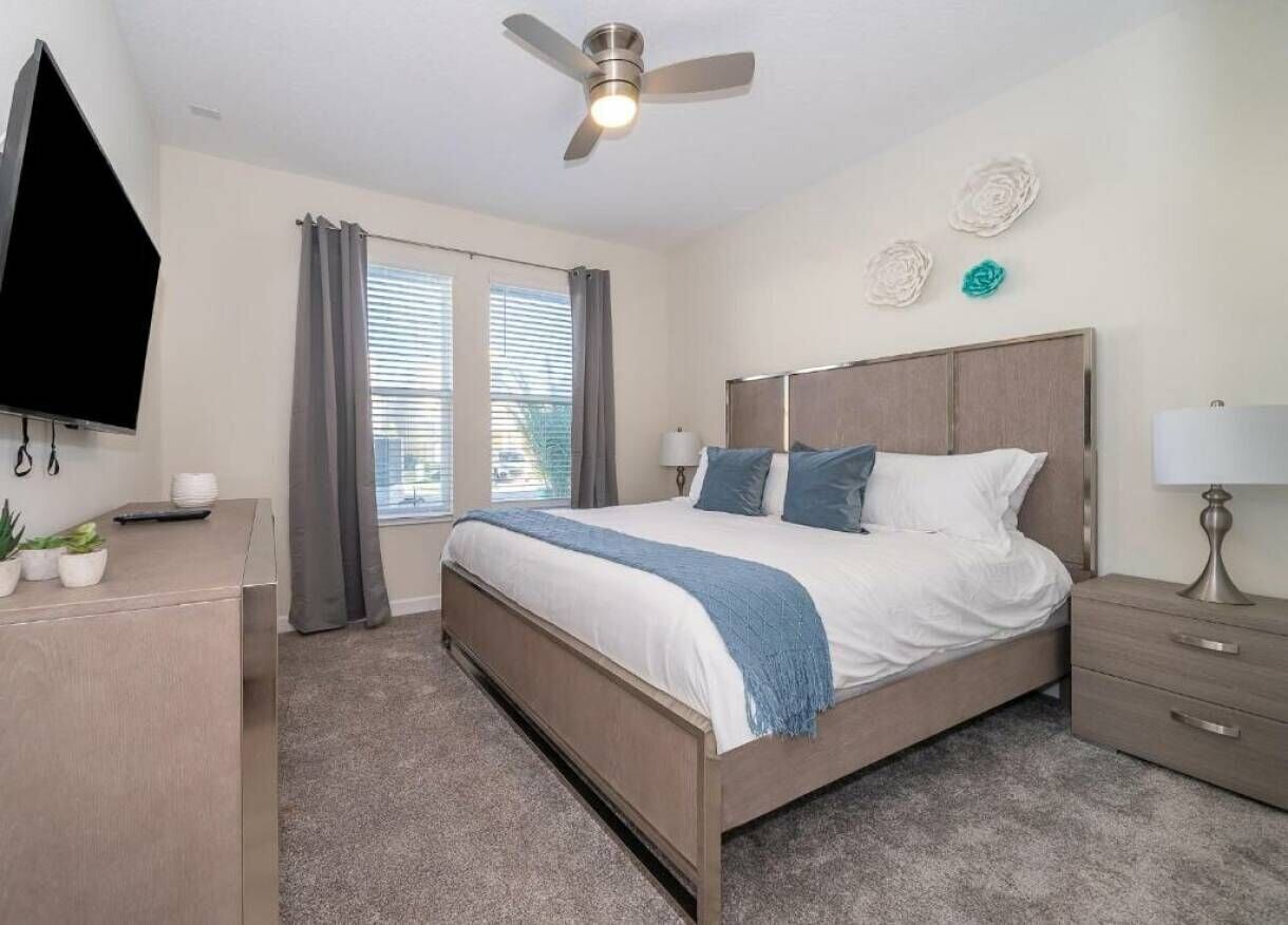 A bedroom with a bed, bedside tables, a ceiling fan, a TV, and a window with curtains.