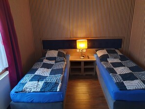 A room with two single beds, a wooden headboard, a nightstand with a lamp, and a window with curtains.