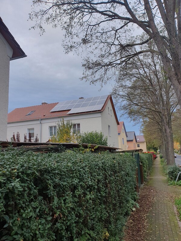 A residential area with a house featuring solar panels on the roof, a paved pathway, and a hedge in the foreground.