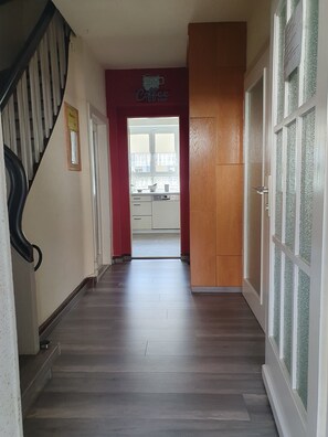 Un pasillo con piso de madera, una escalera a la izquierda y una zona de cocina visible a través de una puerta de vidrio al final.