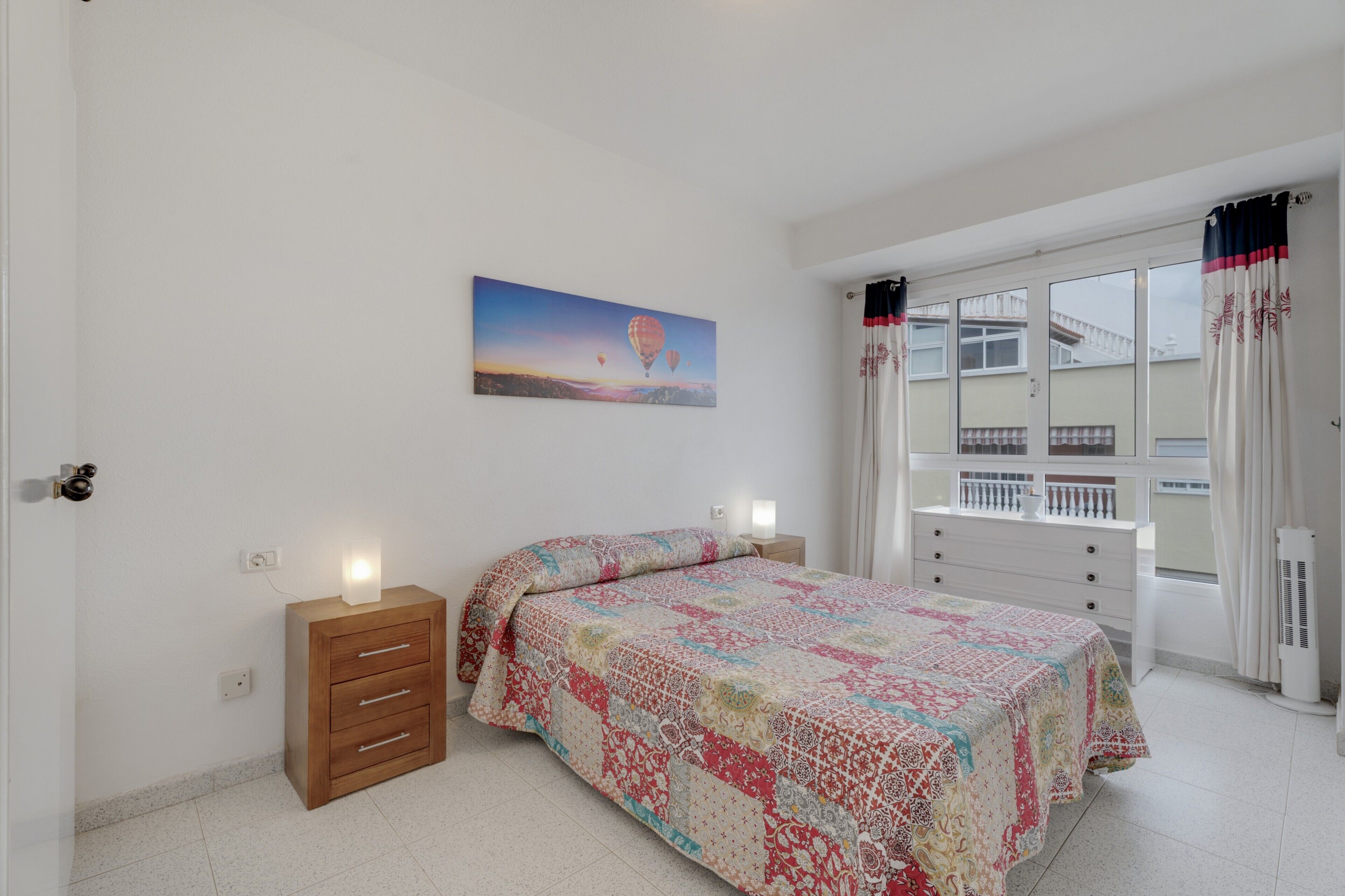 A bedroom with a bed, bedside tables, a window with curtains, a painting on the wall, and a radiator.
