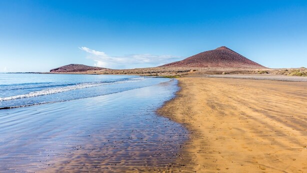 Una playa con dos colinas destacadas, cielo despejado y mar tranquilo.