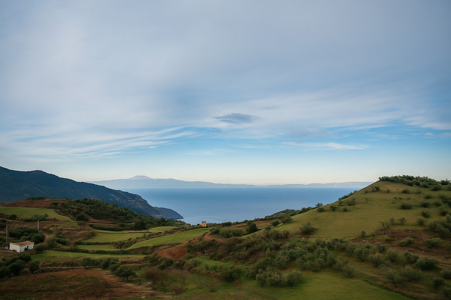 A panoramic view of a coastal landscape with rolling hills, a small building, and distant mountains.