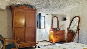A bedroom with a wooden wardrobe, a dresser, a chair, a bed with a floral bedspread, a mirror, and a window with curtains. - Casa Cueva Barranco Hondo (Gáldar)