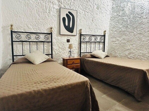 A room with two beds, each with a headboard and a pillow. There is a nightstand with a lamp between the beds. - Casa Cueva Barranco Hondo (Gáldar)