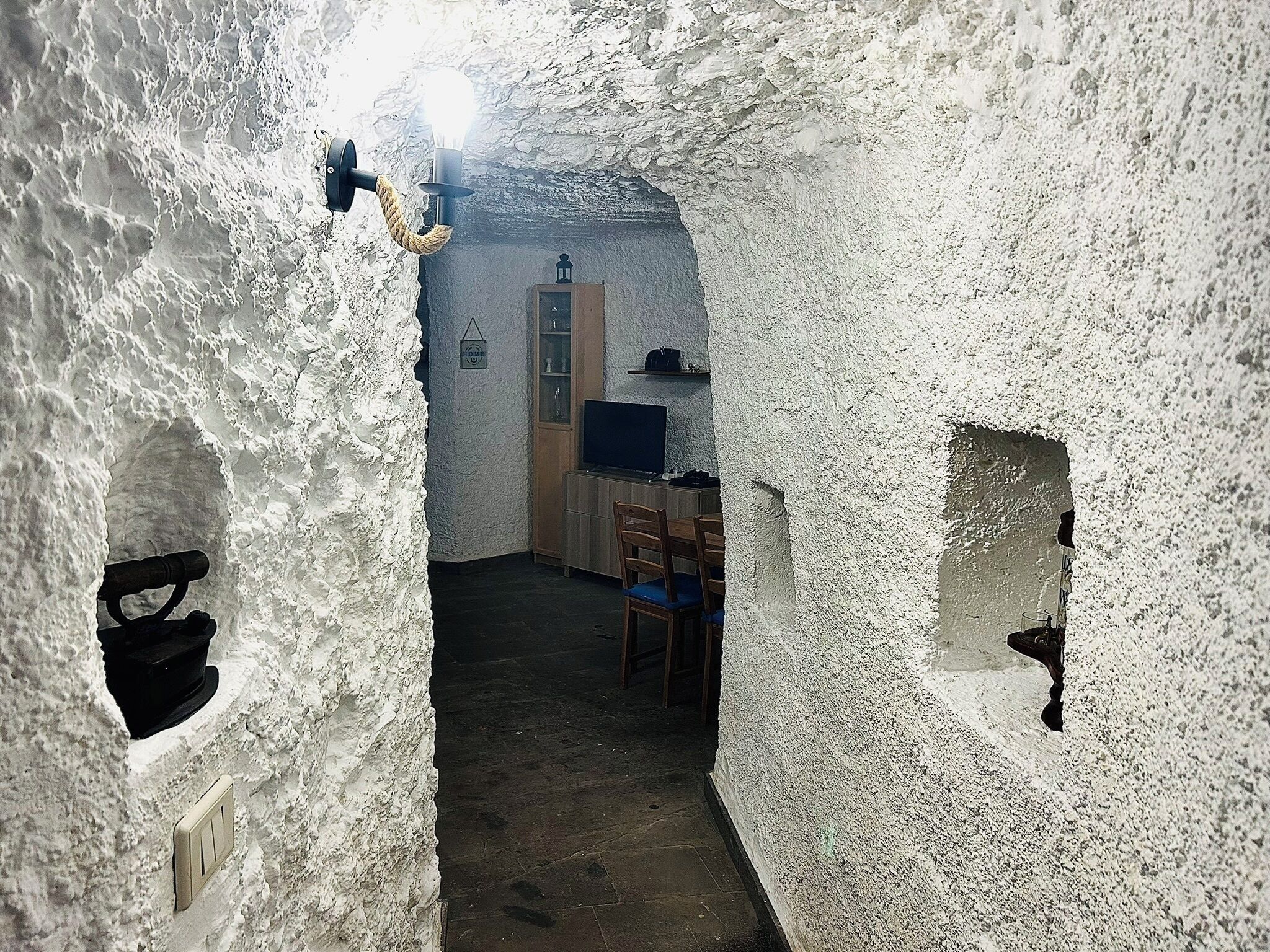A narrow passageway with rough, textured walls, a wooden chair, a television, and a door in the background.