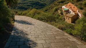 A paved pathway leading to a rustic stone building with a green roof, surrounded by lush greenery and mountains in the background. - Casa Cueva Barranco Hondo (Gáldar)