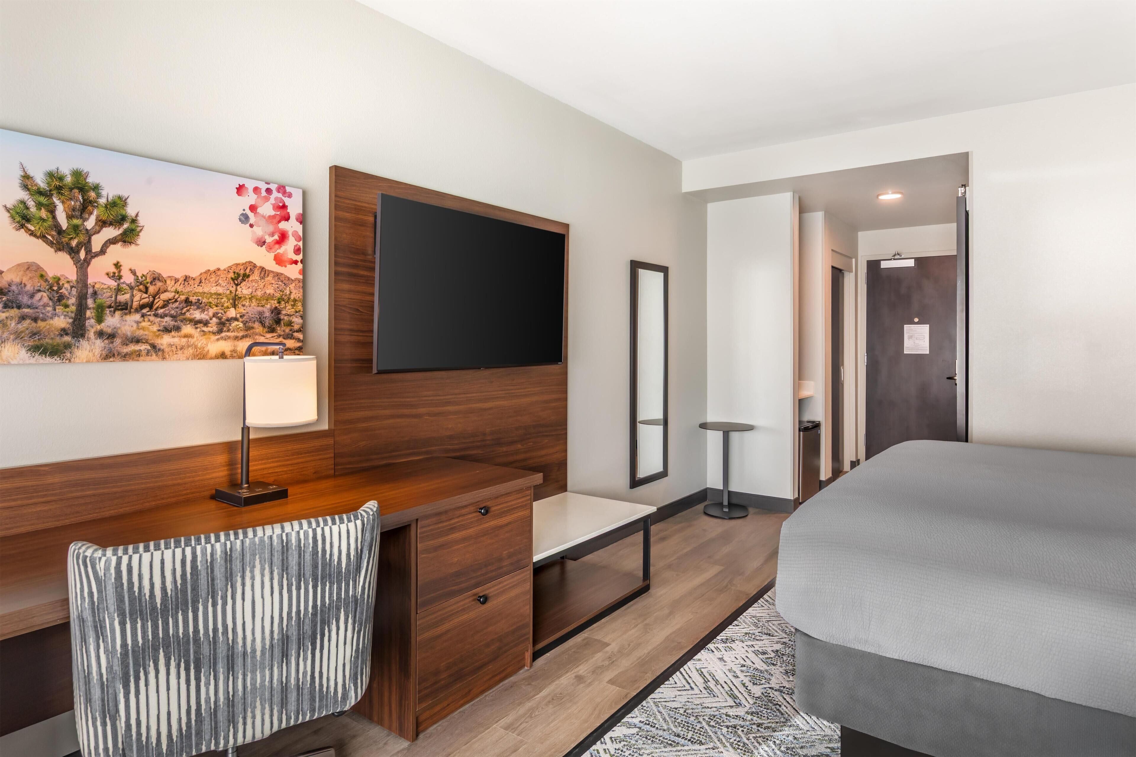 Une chambre d’hôtel moderne avec un bureau en bois, un lit, un téléviseur à écran plat fixé au mur, une lampe de bureau et un tableau représentant un paysage désertique.