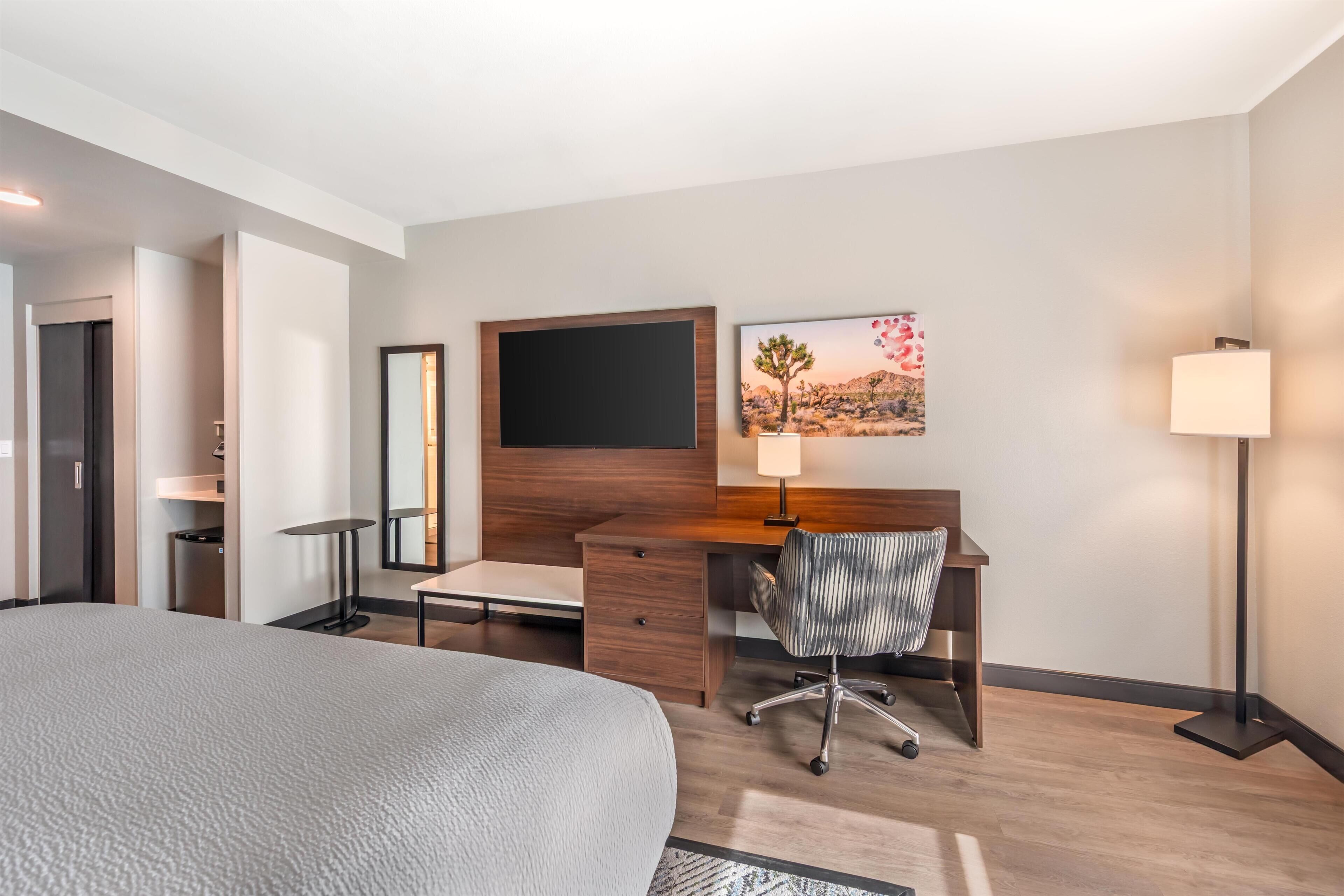 Een hotelkamer met een bed, een bureau met stoel, een televisie, een schilderij en een vloerlamp.