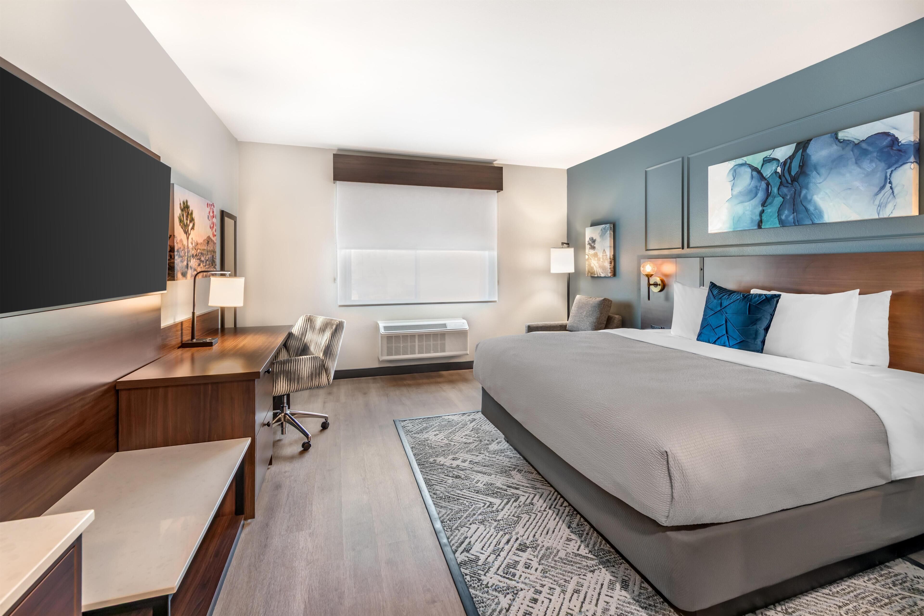 Habitación de hotel con una cama grande, un escritorio, una silla, un televisor de pantalla plana y obras de arte en las paredes.