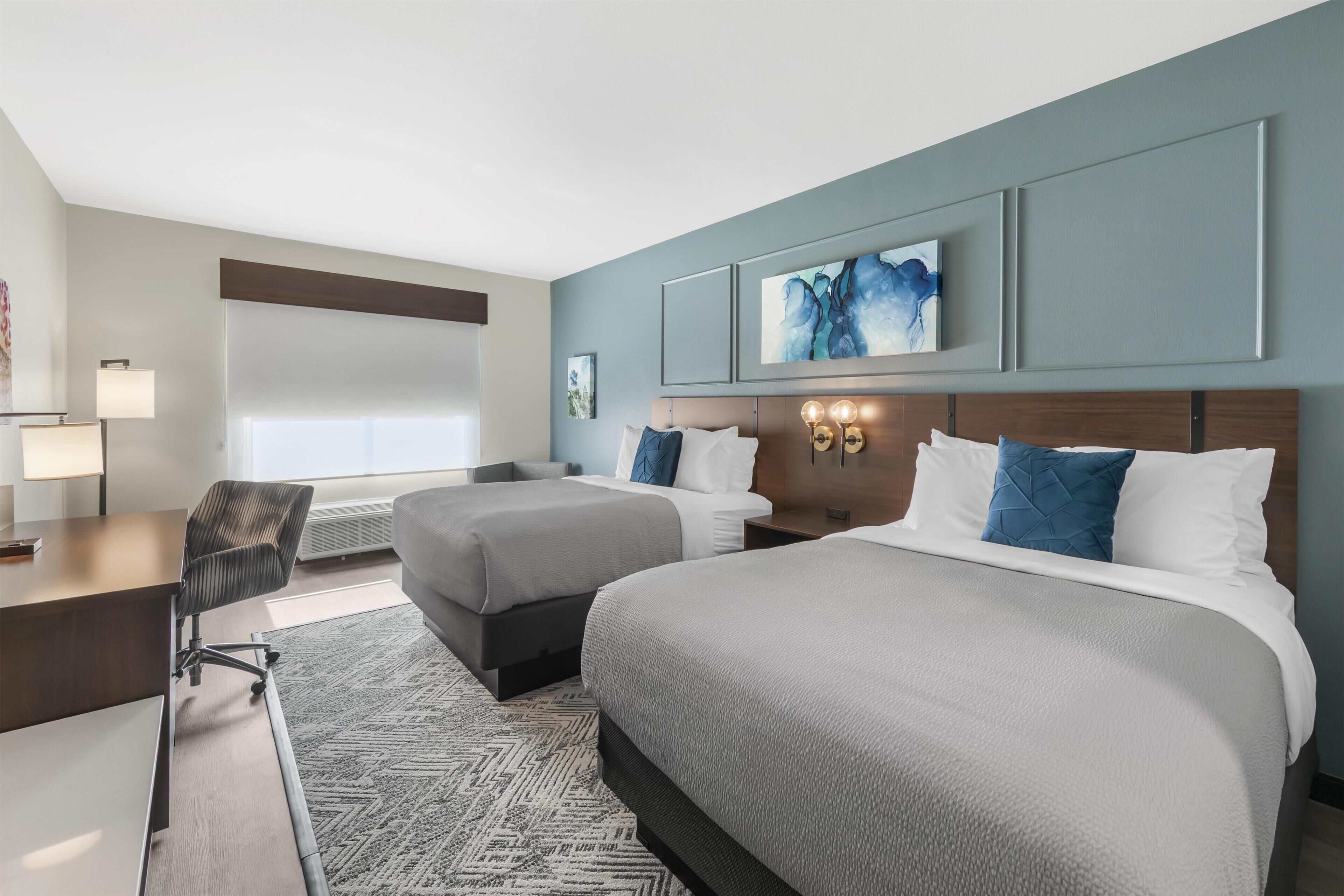 A hotel room with two beds, a desk, a chair, and a large window.