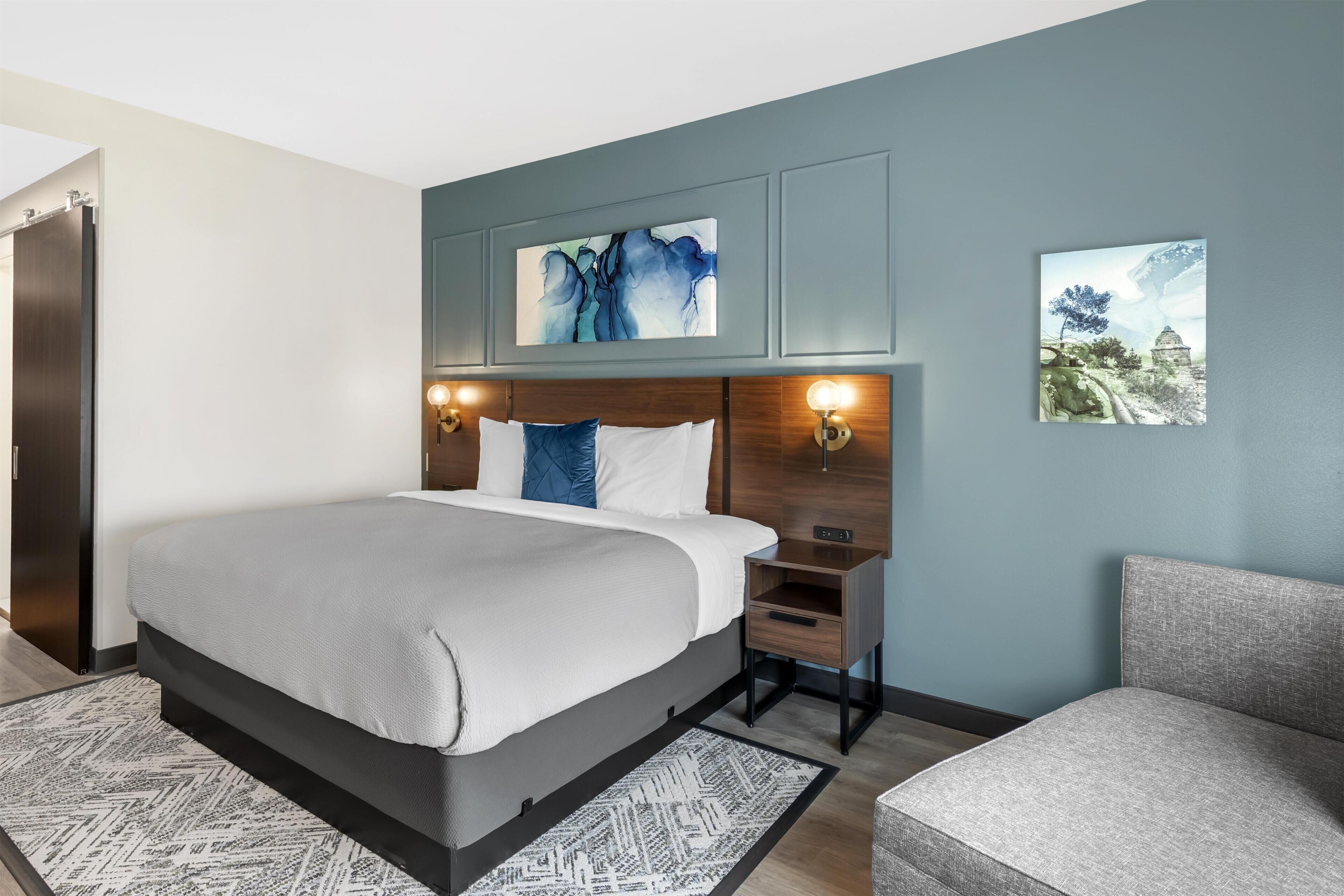 A modern hotel room with a large bed, a nightstand with a lamp, a chair, and artwork on the walls.