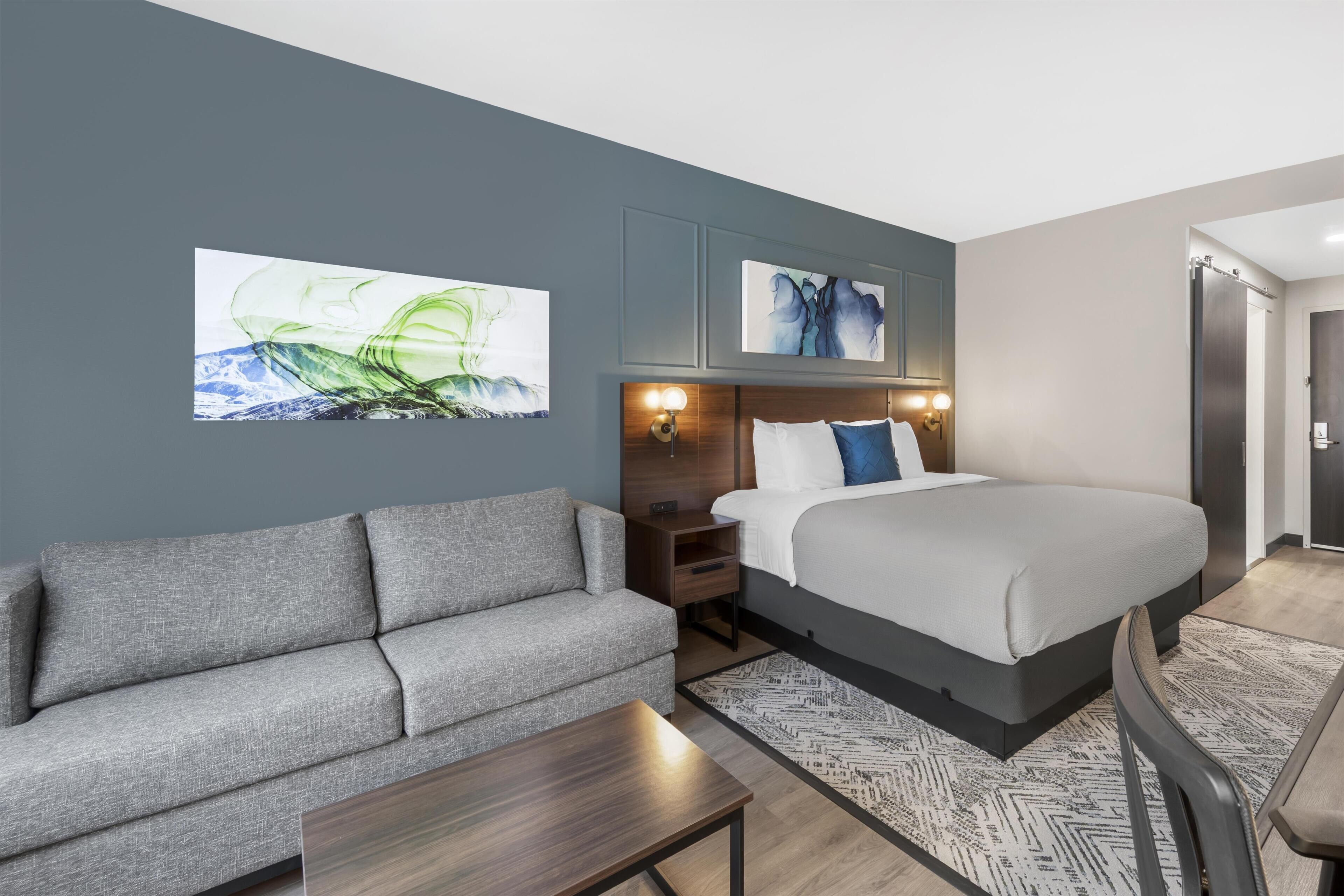 A modern hotel room with a bed, a sofa, a nightstand, and artwork on the walls.