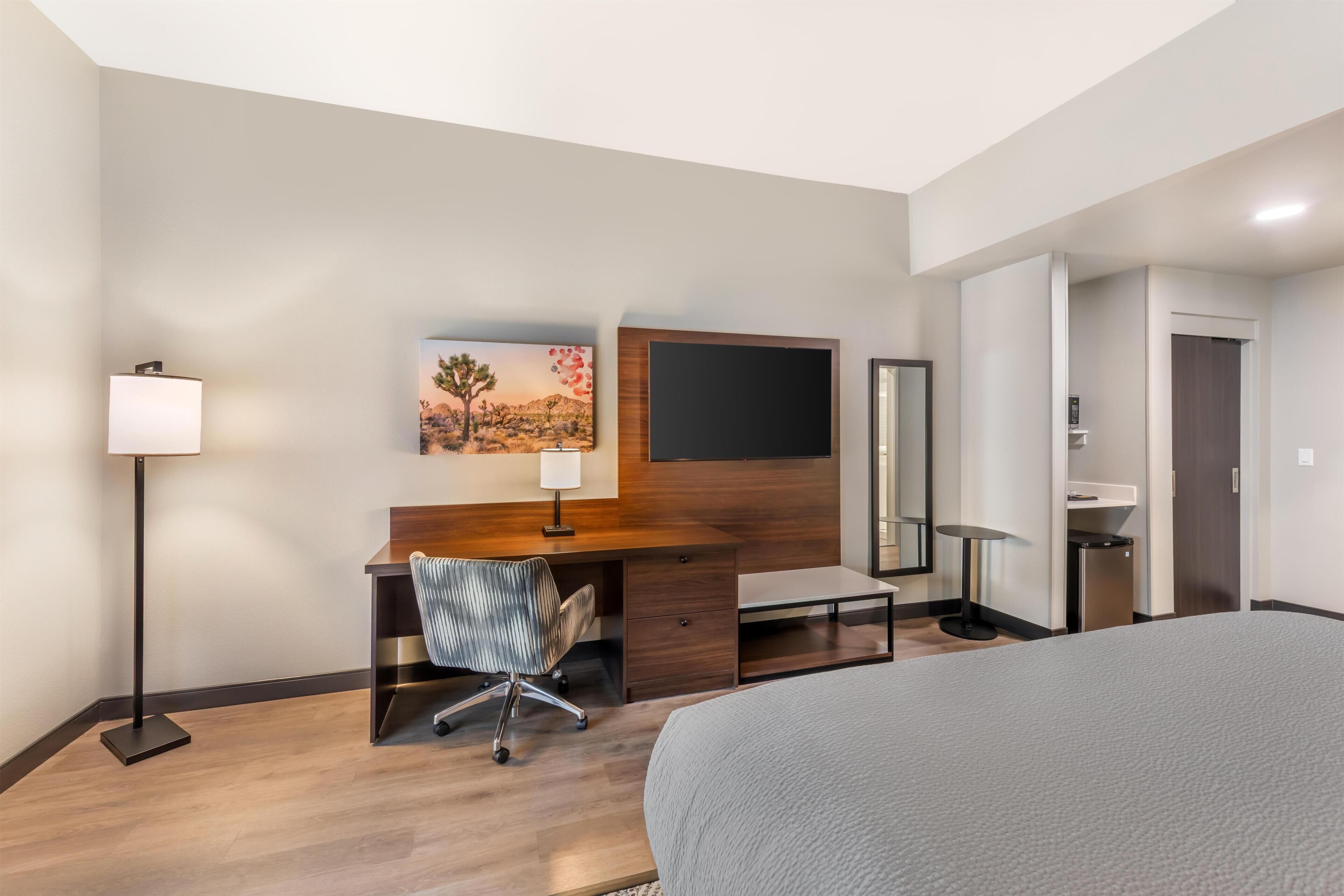 Ein Hotelzimmer mit einem Bett, einem Schreibtisch mit Stuhl, einem Flachbildfernseher, einem Gemälde, einem Spiegel und einer Tür.
