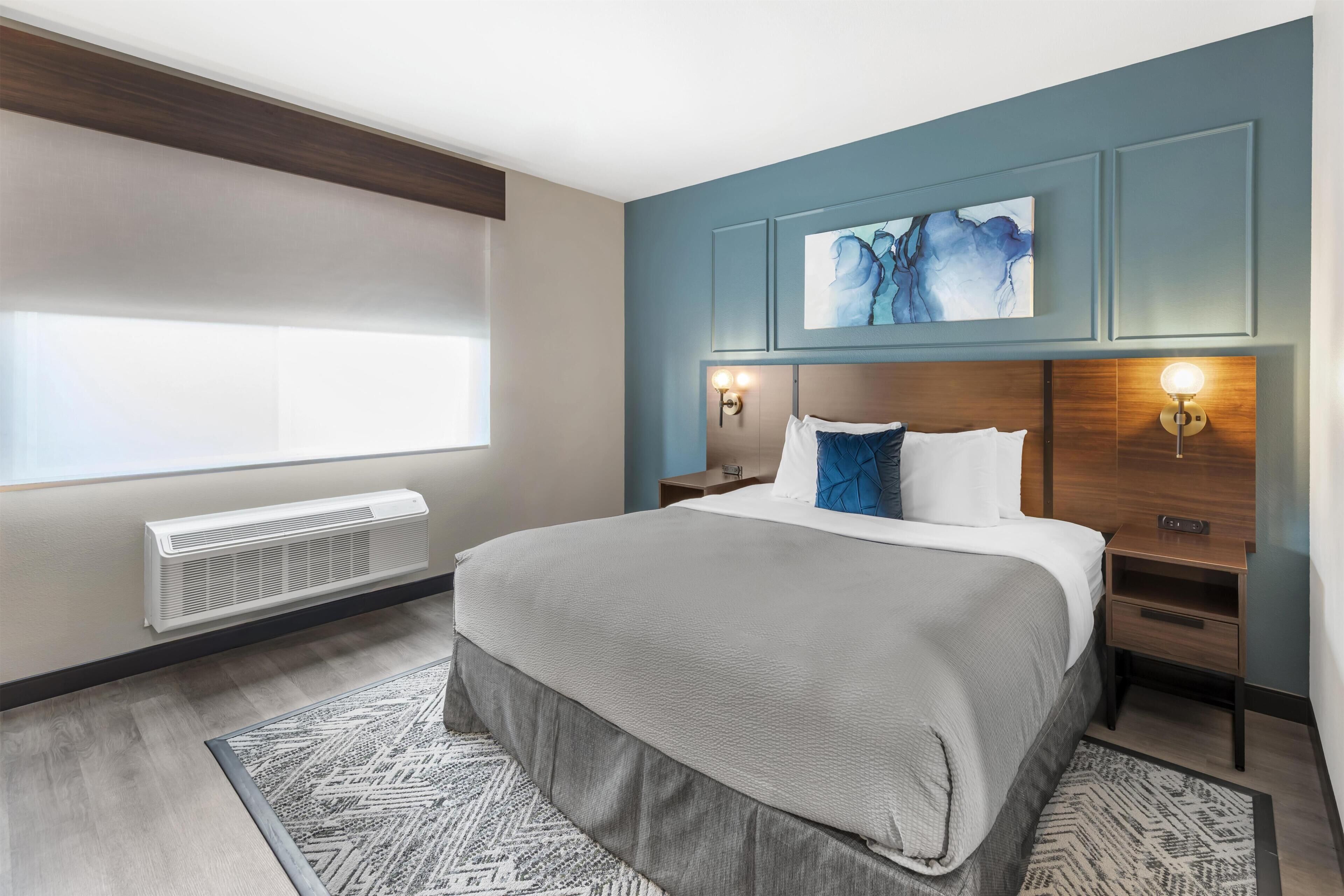 A modern hotel room with a large bed, a wall-mounted artwork, a window with blinds, a heater, and a nightstand with a lamp.