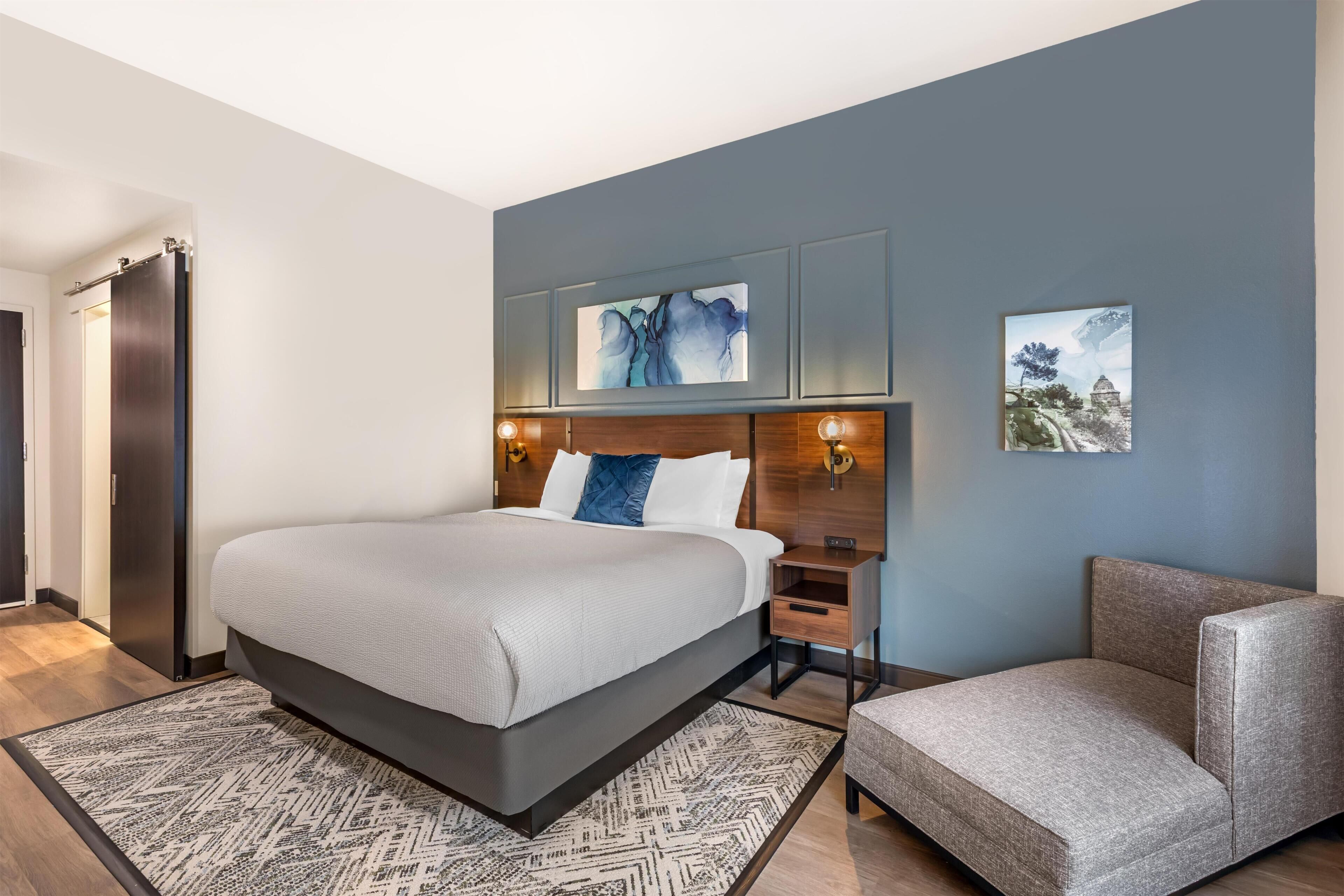 A modern hotel room with a large bed, a bedside table, a chair, and artwork on the walls.