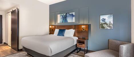 A modern hotel room with a large bed, a bedside table, a chair, and artwork on the walls.