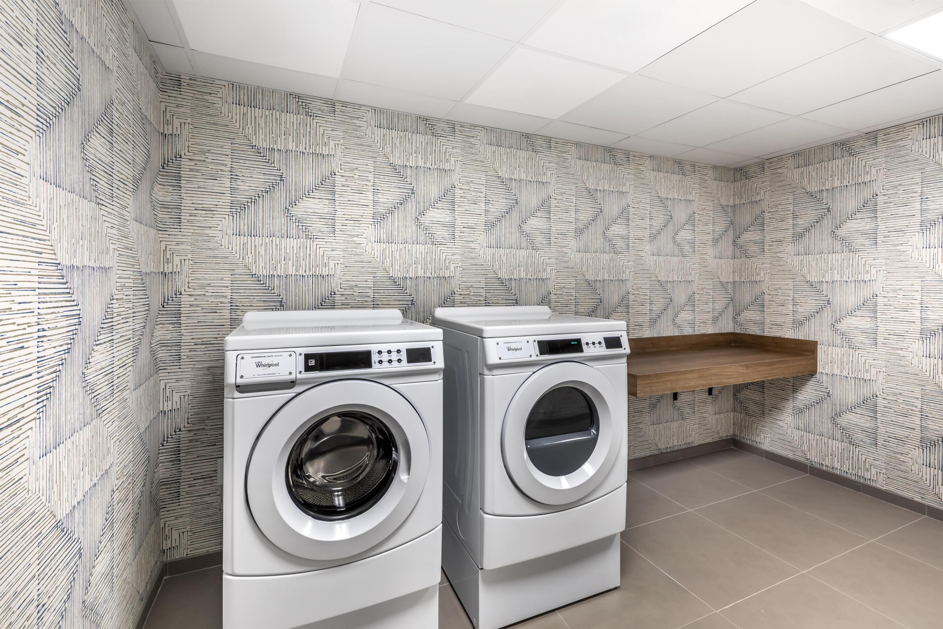 A laundry room with two front-loading washing machines, a wooden shelf, and patterned wallpaper.
