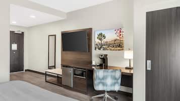 Hotelkamer met een bed, een bureau met stoel, een koffieapparaat, een magnetron, een koelkast, een grote spiegel en een schilderij van een woestijnlandschap.