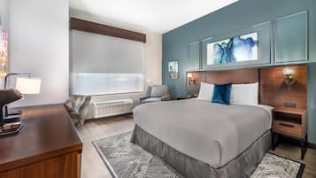 A hotel room with a large bed, a desk with a lamp, a chair, a nightstand, and artwork on the wall.