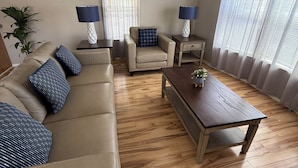 Een woonkamer met een beige bank, een houten salontafel en een stoel met een ruitkussen. Er staan twee lampen op nachtkastjes, een plafondventilator en een raam met dunne gordijnen.
